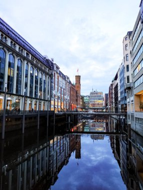 Alacakaranlıkta sessiz bir Hamburg kanalının dikey görüntüsü. Sıcak akşam ışığı binaları yıkıyor ve suyun yüzeyinde yansımalar parıldıyor. Sakin şehir atmosferi.
