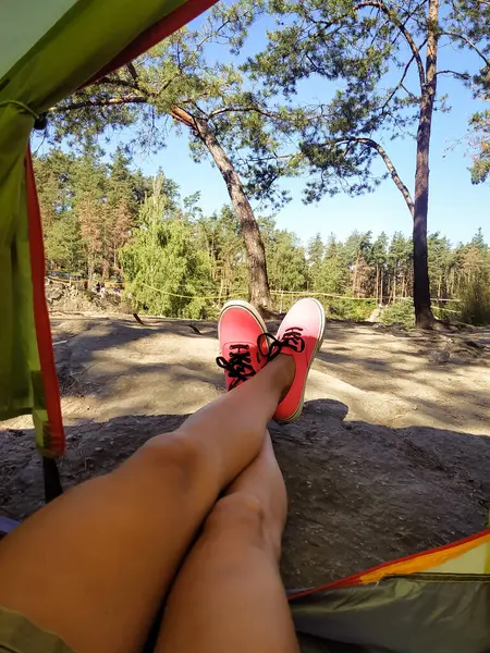 Çam ağaçlarının ve taşların olduğu bir kamp çadırının çıkışında pembe spor ayakkabıların içinde bacaklar. Rahat bir yaşam tarzı, ormanda yürüyüş ve seyahat macerası..