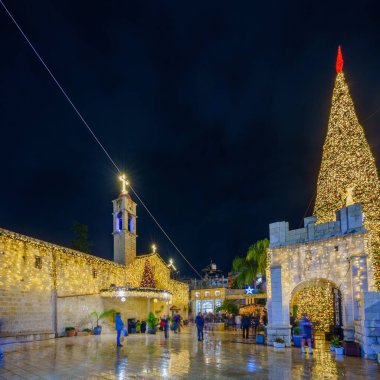 Nazareth, Israel - January 06, 2023: Evening scene of the church (Mary well) square, with Greek Orthodox Church of the Annunciation, Christmas tree, and crowd, Orthodox Christmas Eve, Nazareth, Israel