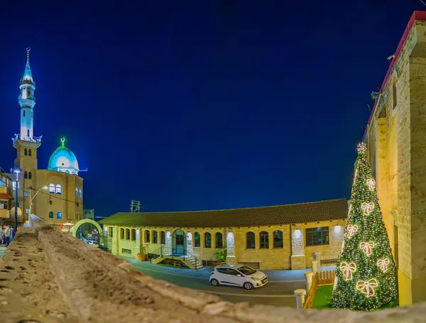 Shefa-Amr, İsrail - 19 Aralık 2024: St. Peter ve St. Paul Kilisesi 'nin bahçesi, bir Noel ağacı ve arka planda Eski Cami. Shefa-Amr (Shfaram), İsrail
