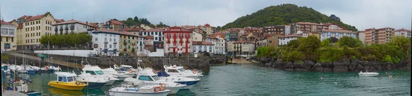 Resimli Mundaka limanının panoramik görüntüsü, renkli evler, botlar ve yağmurlu bir günde arka planda yeşil bir dağ..