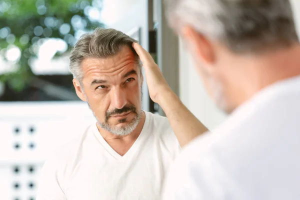 Handsome middle age caucasian man with beard looks in the mirror. Middle-aged man concerned by hair loss. Men\'s personal care. Morning routine. Receding hairline, haircare, beauty concept