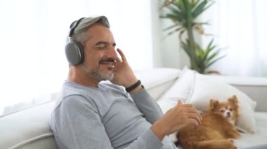 Yakışıklı, orta yaşlı, kablosuz kulaklıklı, en sevdiği müziği dinleyip dans eden sevimli köpeğiyle koltukta oturan beyaz bir adam. Erkek Rahatlaması Koltukta Dinleniyor En sevdiği şarkının tadını çıkarıyor.