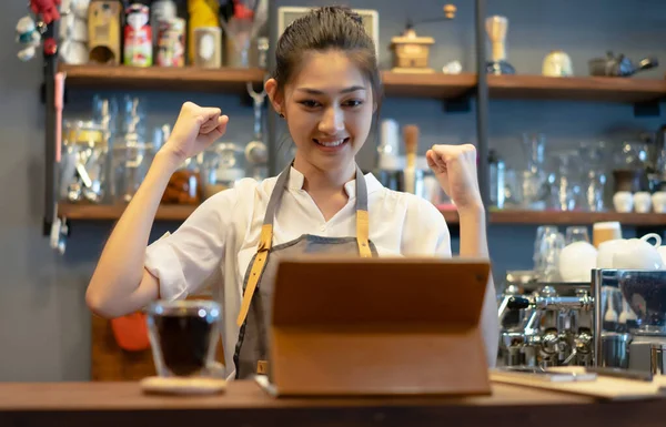 Encouraged happy young Asian woman barista lift hands up and shouting yes while looking at digital tablet with large quantities of orders. Celebrate success or win. Small Business and Owner Concept
