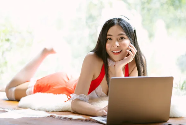 Portrait of beautiful sexy Asian woman lying on carpet with laptop computer to surf the internet with a happy relaxed smile and looking at camera in living room. Enjoy free and Relax time in the house
