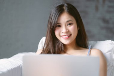 Beautiful Asian woman using notebook digital watching video online, chatting with friends and looking at camera while sit on couch in living room at home. Cheerful Korean girl enjoy weekend free time.