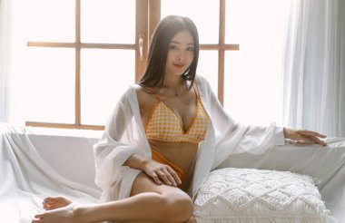 Portrait of beautiful sexy Asian woman in orange bikini posing and looking at camera while sitting on sofa in living room. People lifestyle, Female beauty, spa concept