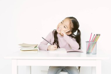 Portrait of little kid Asian schoolgirl sitting at desk handwrite in notebook having distant class, Asia kid prepare homework study alone isolated on white wall background. Homeschooling concept