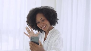 Afro-Amerikan güzel bir kadının portresi gülümseyen neşeli bir güzel çevrimiçi video çağrısı yayınlıyor blogcularla tanışıyor ve yatak odasında otururken akıllı telefondan merhaba diyor..