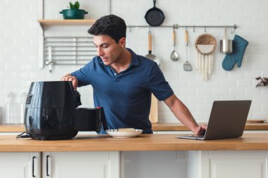 Yakışıklı adam mutfak masasında duruyor ve ekmek pişiriyor. Air Fryer makinesinin tarifiyle sağlıklı. Evde modern mutfakta dizüstü bilgisayarla yemek pişirme dersi alıyor..