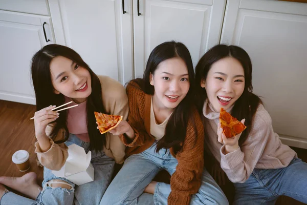 Three young beautiful Asian smiling hipster female sit on the floor and laugh at funny joke eating pizza, takeaway food in modern kitchen at home. Happy korea women having fun and looking at camera.