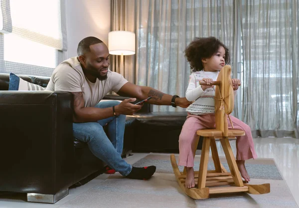 Family enjoy funny moments at home. African American father and daughter playing rocking horse in living room. Happy Black family activity together. Weekend lifestyle concept.
