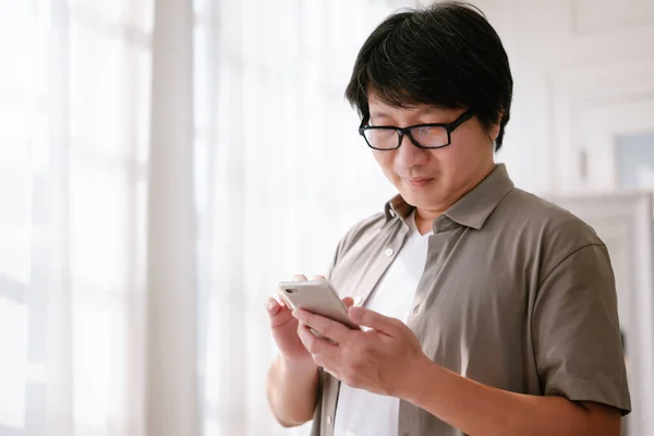 stock image Portrait of Mid adult Asian man wearing casual man with smartphone, working remotely or video chatting in living room. Middle Korean man browsing internet, having online meeting, using app at home.