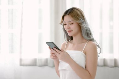 Güzel, genç Asyalı bir kadın ayakta duruyor ve mesaj yazıyor, cep telefonuyla sosyal medyayla sohbet ediyor, evdeki oturma odasında online iletişimden hoşlanıyor. Teknoloji iletişim yaşam tarzları konsepti.