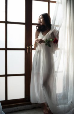 Mükemmel ince yapılı vücut portresi güzel yüz zarif Asyalı kadın kırmızı güllü beyaz elbise giyiyor ve dışarıdaki pencereye bakıyor. Moda ve güzellik konsepti.