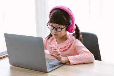 Kulaklık takan küçük Asyalı kız dizüstü bilgisayarını kullanarak online bir ders veriyor. Bilgisayarda bilgisayar başında bilgisayar başında bilgisayar ekranına bakıp, internet üzerinden ödev yapan mutlu bir öğrenci..