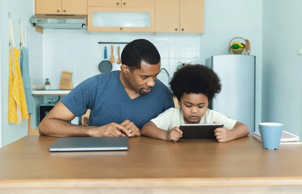 Happy African American father and his little son using digital tablet sits together at table in kitchen room. Caring dad and small boy child do home task learn learn online clase. Education concept.