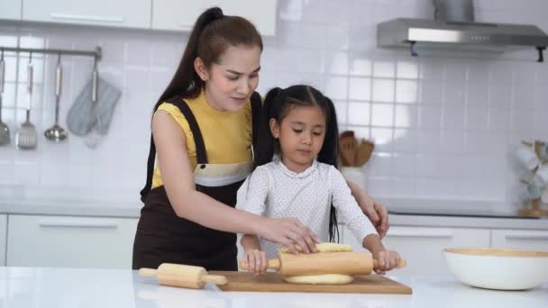 ハッピーアジアの家族が一緒にパン屋を準備しています アジアの母は彼女の小さな娘にキッチンで一緒に焼いている間に生地をひざまずく方法を教えています 自家製の食べ物と小さなヘルパー — ストック動画