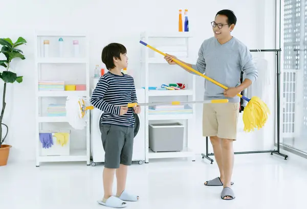 Happy Asian family cleaning house and having fun. Father dancing and singing with son cleaning the laundry room together. Smiling single father pretend to play music holding mops like guitars at home