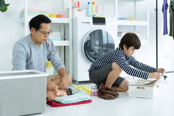 Asian father and kid little boy spends time together in the laundry room. Single father and his son sitting the floor helping of sorting clothes, folding laundered items. Home cleaning concept.