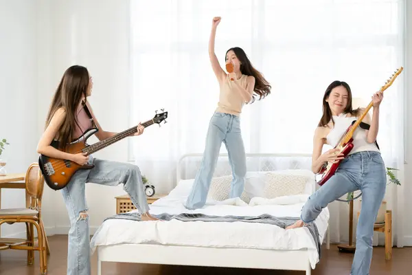 Happy group of Asian friends singing and jumping on bed at home. Cheerful Asia women have fun together playing guitar with melody singing hairbrush as microphone. Lifestyle, Hobby, Friendship concept