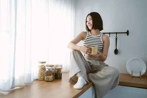 Portrait of beautiful Asian woman in cozy clothes relax sitting on counter top at kitchen drinking coffee and looking through window at home. Spending free time cozy at home. Emotion Positive Concept