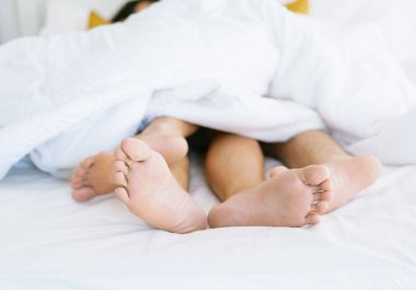 Cropped image of bare feet of loving gay couple lying under white blanket in cozy bed at home against blurred background