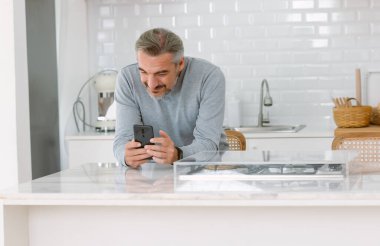 Modern mutfak adasında dikilirken cep telefonu kullanan yakışıklı, beyaz sakallı yaşlı adamın portresi. Tatil etkinliği ve İletişim Teknolojisi Konsepti.