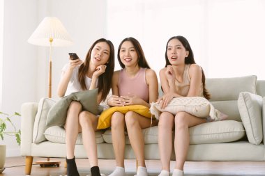Kanepede oturup televizyon izleyen, uzaktan kumandalı, gülümseyen ve gülen, evdeki oturma odasında rahatlık ve sıcaklık hisseden üç güzel Asyalı kadın..
