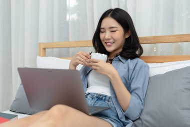 Yatak odasında sıcak kahve içip dizüstü bilgisayar kullanan Asyalı güzel bir kadının gülümsemesi. Özgür ve mutlu anların tadını çıkarıyorum. Teknoloji ve sosyal medya kavramı