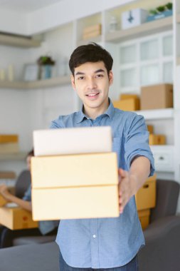 Arka planda çalışan bir sürü karton kartonla mutlu bir Asyalı genç adam. Neşeli erkek, teslimat servisinden bir sürü paket alıyor. Serbest çalışan.