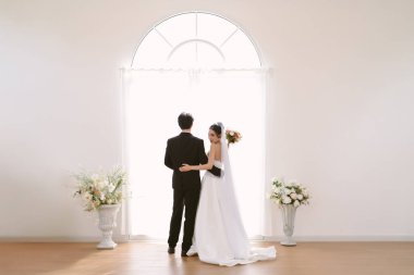 Asyalı güzel gelin birbirine sarılıyor ve pencereli beyaz giriş kapısına bir buket çiçek atmaya hazırlanıyor. Genç ve güzel kadın ve erkek takım elbiseli ve kısa elbiseli..