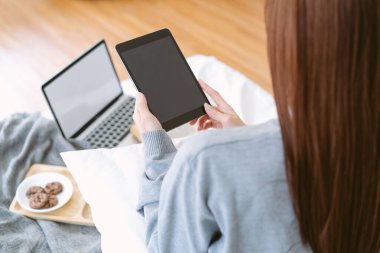 Yanında laptoplar, kurabiyeler ve kahveyle yatakta otururken gri kazaklı Asyalı kadının arka görüntüsü. Dijital hayatı, rahatlığı ve modern evi gösteren rahat bir yaşam tarzı..