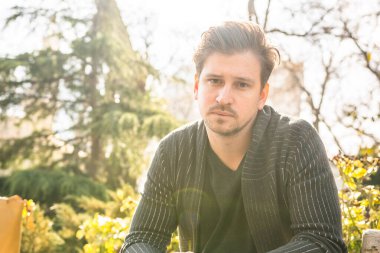 Parkta güneş ışığında siyah ceketli, siyah gömlekli bir adam oturuyor.