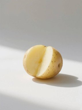 Açık sarı etli yarım bir patates ve küçük kahverengi benekli ince, soluk sarı bir ten. Sağında yumuşak bir gölge olan beyaz bir yüzeyde oturuyor..