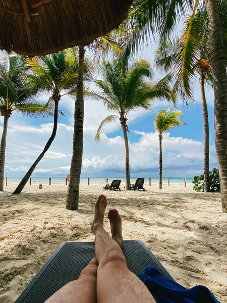 Karayipler sahilinde, plaj sandalyeleri, sazdan şemsiyeler ve palmiye ağaçlarıyla dinlenen insan bacakları. Playa del Carmen, Meksika 'da yaz tatili.