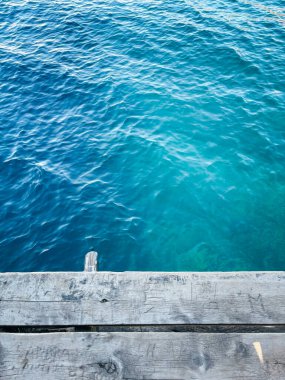 A weathered wooden dock extends over clear turquoise water, with gentle ripples on the surface reflecting sunlight, creating a tranquil and serene lakeside view. clipart