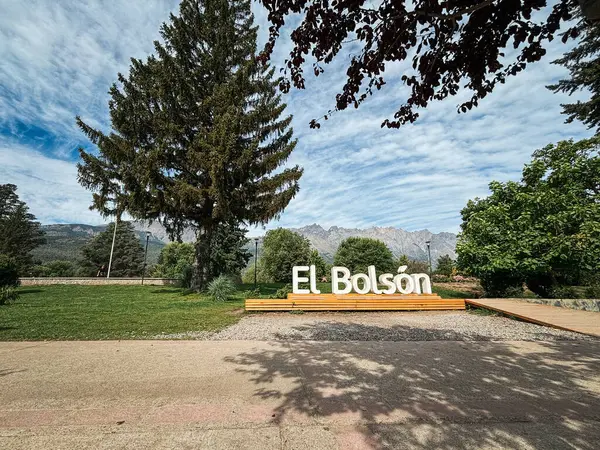 Parktaki El Bolson tabelasının manzarası. Etrafı yemyeşil, yüksek ağaçlarla çevrili. Arka planda ise bulutlu bir gökyüzünün altında bir dağ sırası..