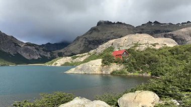 Turkuaz bir göl ve kıyı şeridine yakın parçalı bulutlu bir gökyüzünün altında yuvalanmış kırmızı çatılı ahşap bir kulübe ile ormanlık bir vadinin üzerinde görkemli kayalık tepeler yükselir. Refugio San Martin - Jakob, Bariloche, Arjantin