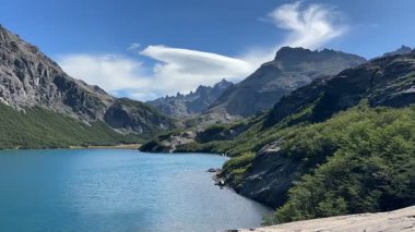 Arjantin 'in Bariloche şehrinde berrak bir gökyüzünün altında yükselen pürüzsüz tepeleri olan orman yamaçları ve kayalık dağlarla çevrili canlı mavi bir göl..
