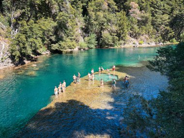İnsanlar sık orman ve kayalık kıyılarla çevrili sakin bir zümrüt yeşili nehirde yüzüyor ve yürüyor. Uçurumlar ve parlak mavi bir gökyüzü verimli bir yaz manzarası yaratıyor.