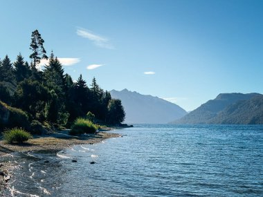 Ağaçlarla ve çalılarla kaplı sakin bir göl kıyısı uzak dağ sırtlarına, berrak gökyüzünün altına ve yumuşak göl yüzeyine sabahın erken saatlerinde yansıyan ışığa götürür.