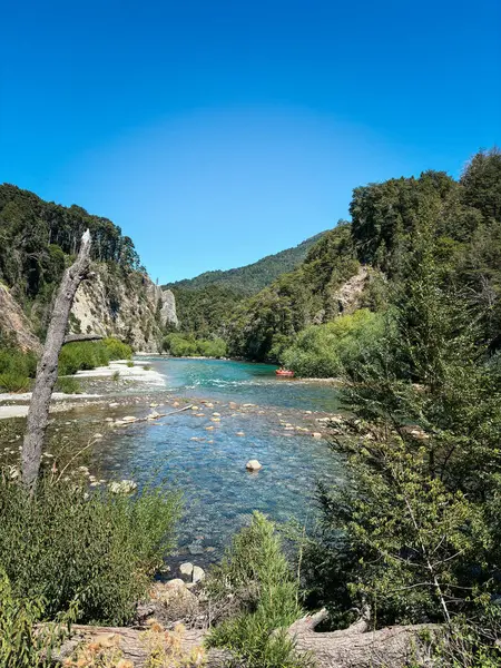 Yeşil ormanlık uçurumlar ve parlak mavi bir gökyüzü arasında uzanan, görülebilir taşlarla dolu el değmemiş bir nehir. Uzakta yüzen küçük kırmızı bir sal.