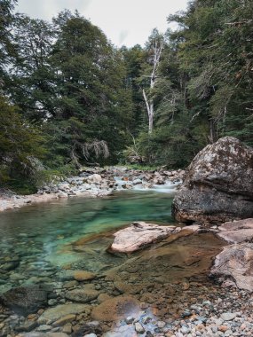 El değmemiş turkuaz bir nehir, yoğun yeşil ormanlarla ve bulutlu gökyüzünün altındaki büyük kayalarla çevrili kayaların ve çakıl taşlarının üzerinden nazikçe akar.