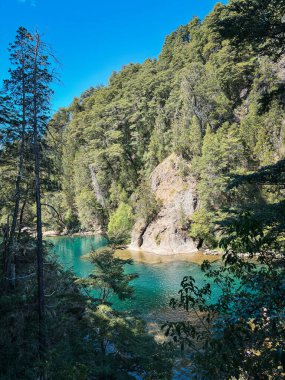 Yapraklı ağaçlar ve sık ormanlardan oluşan bir çerçevenin içinden görünen gizli bir turkuaz nehir berrak mavi gökyüzünün altında sakin sudan yansıyan güneş ışığıyla görülebilir.