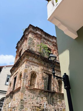 Tarihi bir binanın tuğla ve taş kalıntıları, muhtemelen bir kilise, tepeden büyüyen bitkiler. Klasik bir sokak lambası ön planda duruyor, eski mimariyle şehir hayatının çelişkisi.