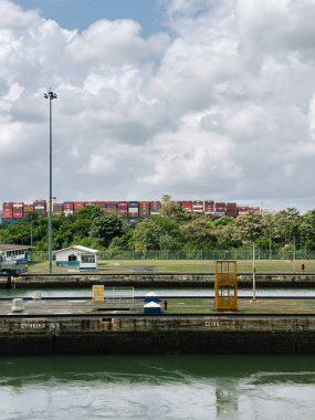 Panama Kanalı 'nın geniş bir görüntüsü, beton bir su yolu, beyaz bir bina ve ahşap bir saat kulesi. Çok renkli kargo konteynırlarının büyük bir yığını ağaç hattının üzerindeki bir gemide görülebilir.
