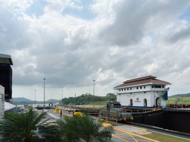 Miraflores Kilitleri 'nin beyaz ziyaretçi merkezi binasını ve kilit operasyonlarını gösteren geniş bir görüntüsü. Ön planda yeşil palmiye ağaçları, su manzaralı ve bulutlu bir gökyüzünün altında tropik tepeler vardır.