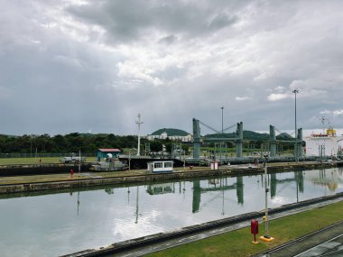 Panama Kanalı 'nda bulutlu bir günde büyük bir kargo gemisinin tutulduğu geniş bir manzara. Su durgun, manzara yemyeşil ve yemyeşil, kanal boyunca altyapı görünür.