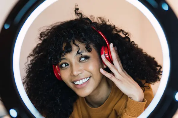 Happy young African American latina woman listening to music using white headphones at home bedroom.